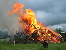 Ein brennender Holzhaufen auf einem Feld. Im Feuer sind Möbelstücke und EPAL-Paletten zu erkennen, eine braune Rauchfahne steigt auf. Vor dem Feuer sitzt ein Kind.